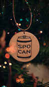 Decorative leather ornament with aluminum can design and 'SPO CAN' text hanging on a Christmas tree with blurred lights in the background.