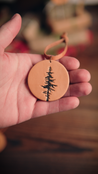 Hand holding a handcrafted leather ornament with a Lodgepole Pine Tree design against a blurred background