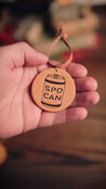 Hand holding a handcrafted leather ornament with an aluminum can design and text 'SPO CAN' against a blurred background