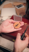 Hand holding a leather ornament with a pine tree engraving, surrounded by tools and materials.